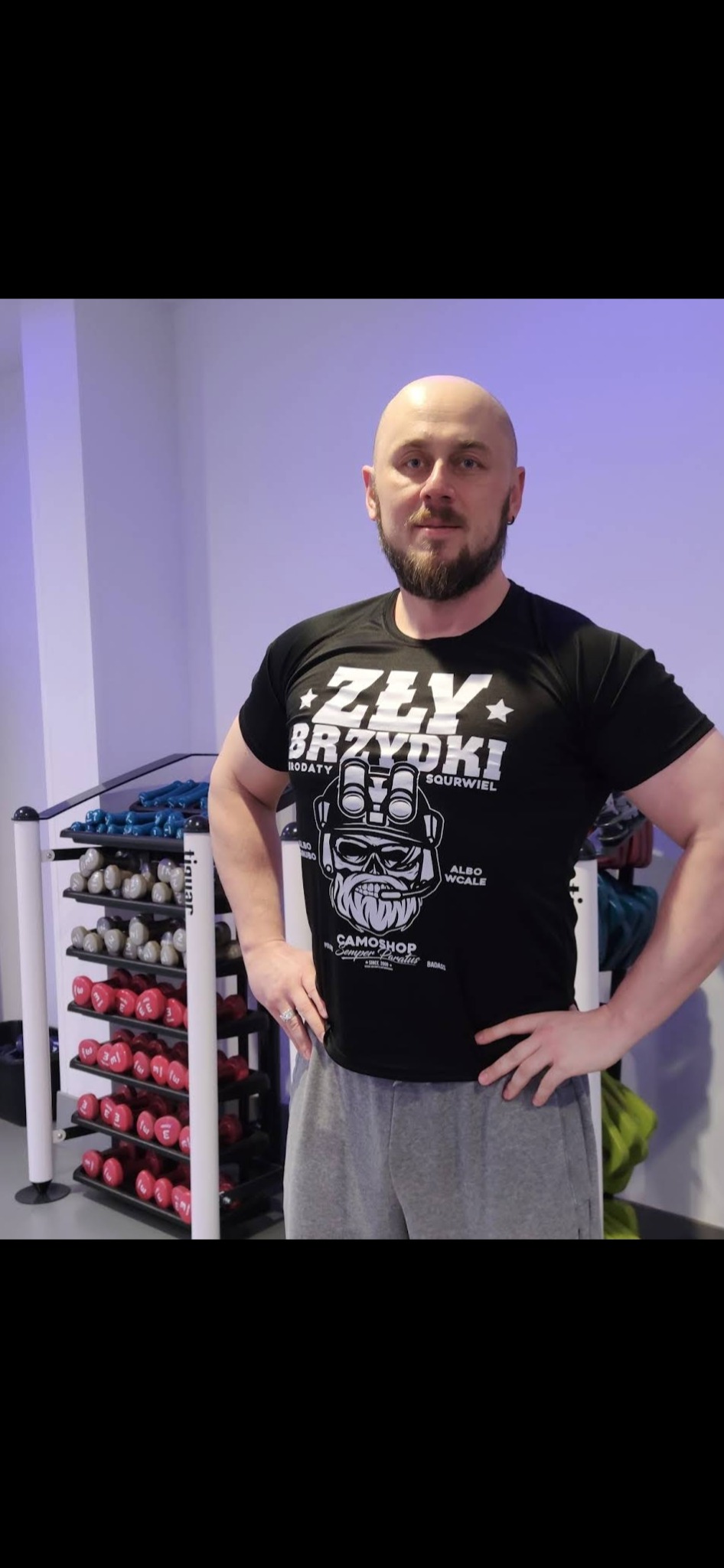 Mężczyzna w czarnej koszulce z nadrukiem, stojący w siłowni na tle regału z hantlami.