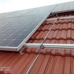 Instal Volt - Panele słoneczne zamontowane na dachu z czerwonej dachówki, widoczne aluminiowe profile montażowe i okablowanie, pochmurne niebo w tle.