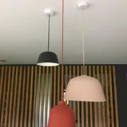 Trzy nowoczesne lampy wiszące w kolorach: czarnym, pomarańczowym i pudrowym różu, na tle drewnianej ścianki działowej.