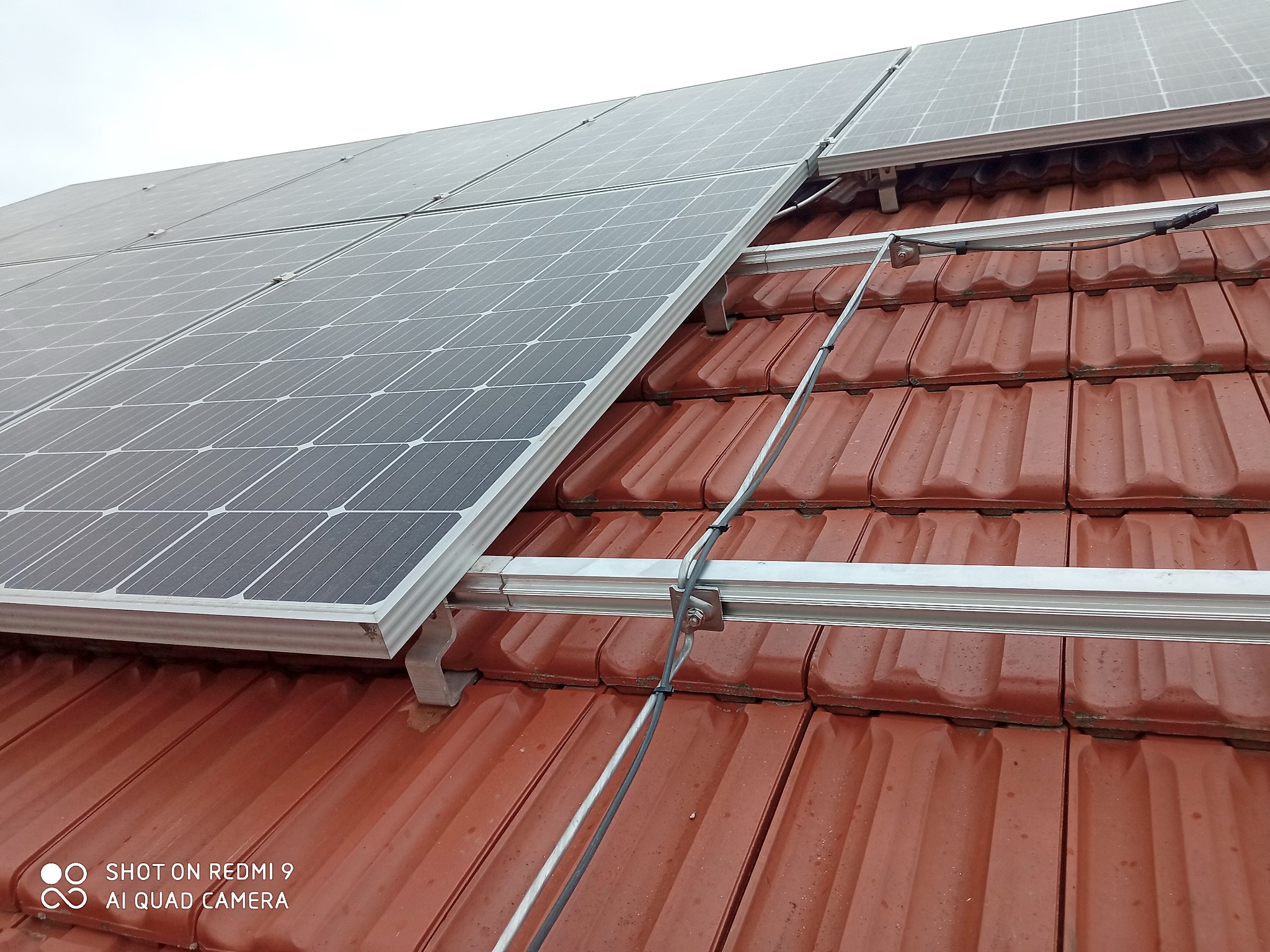 Panele słoneczne zamontowane na dachu z czerwonej dachówki, widoczne aluminiowe profile montażowe i okablowanie, pochmurne niebo w tle.