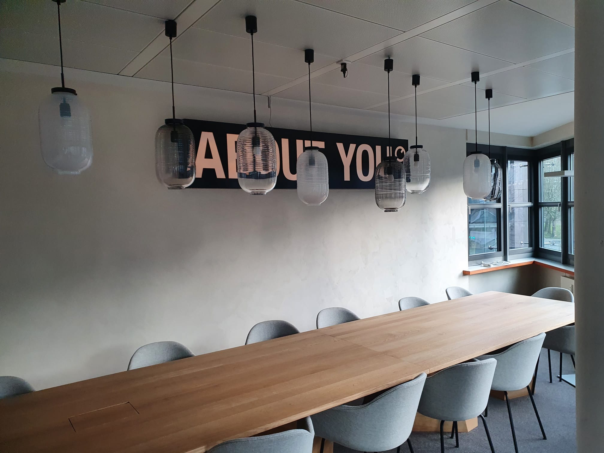 Sala konferencyjna z długim drewnianym stołem otoczonym szarymi krzesłami, oświetlona wiszącymi lampami o szklanych kloszach, na ścianie napis 'ABOUT YOU' pod sufitem z widocznym systemem oświetlenia.