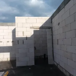 Wnętrze w trakcie budowy z białych bloczków, widoczne ściany z otworami drzwiowymi, szare bloczki na górze, wilgotna podłoga i pochmurne niebo w tle.
