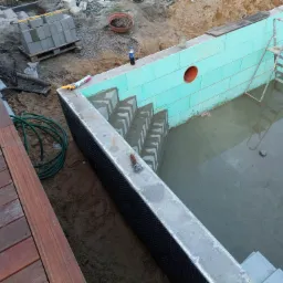 Budowa basenu: ściany z zielonych bloczków izolacyjnych, betonowe schody wewnątrz, widoczna drewniana podłoga tarasu i materiały budowlane wokół.