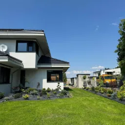 Nowoczesny dom z zagospodarowanym ogrodem: świeżo posiana trawa, rabaty z krzewami iglastymi i ozdobnymi, czarna agrowłóknina, żółty samochód dostawczy w tle.