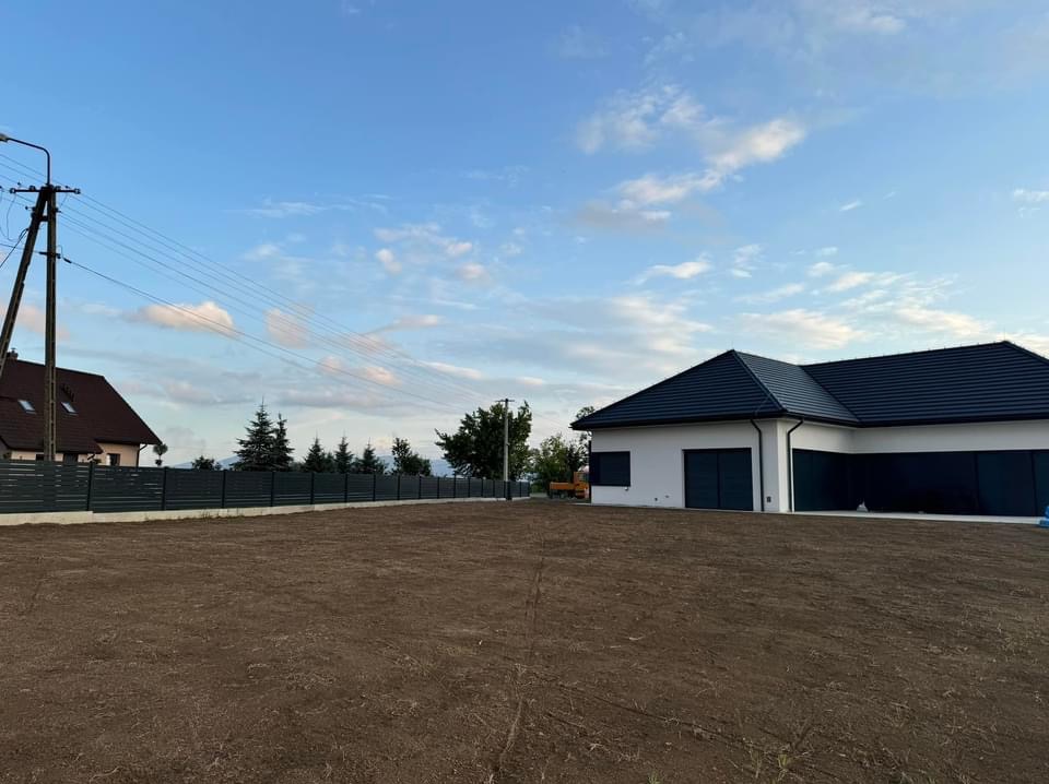 Przygotowany teren pod trawnik z widocznym budynkiem mieszkalnym i ogrodzeniem panelowym w tle.