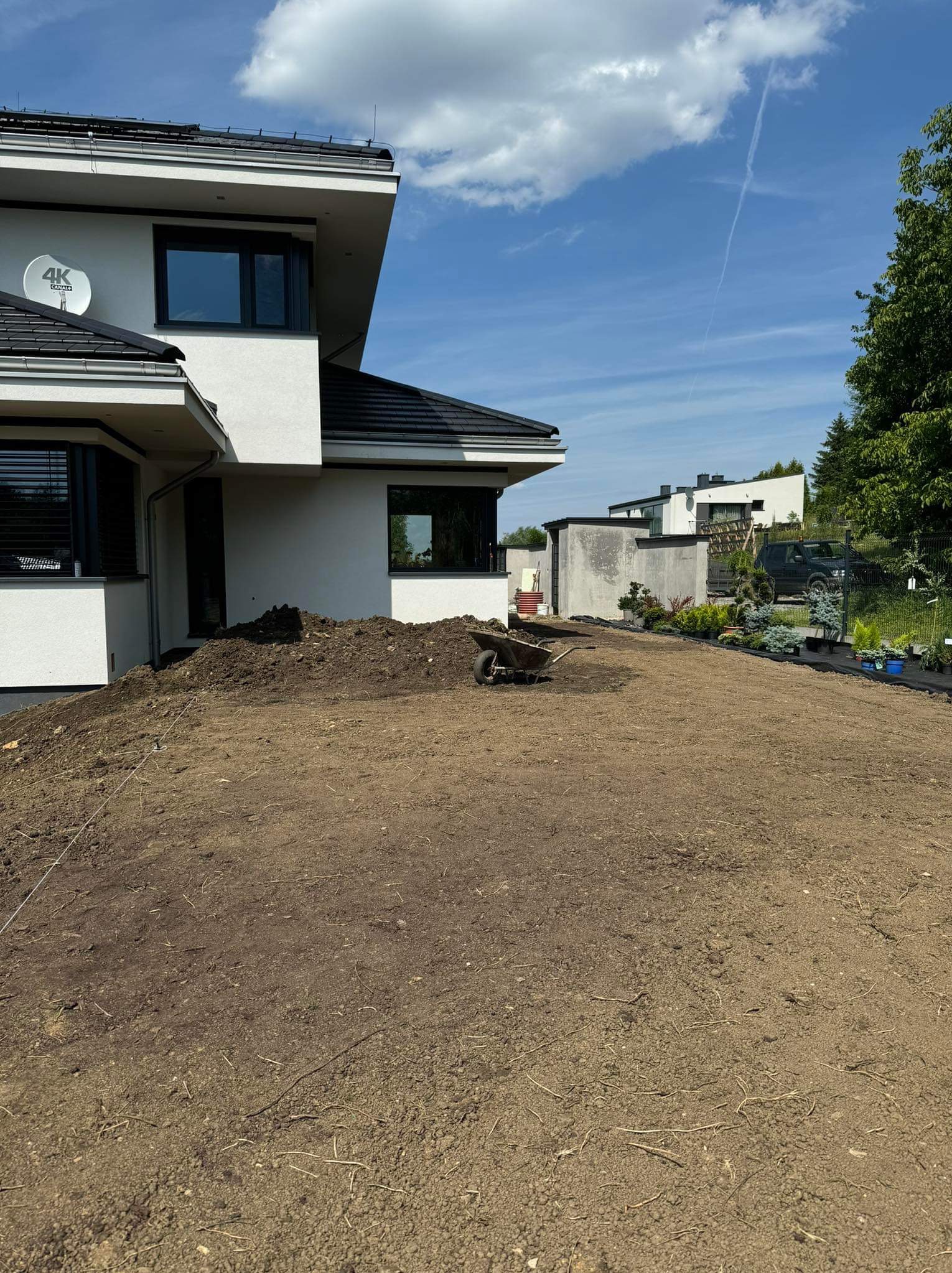 Przygotowany teren pod trawnik przy nowoczesnym domu, z widoczną taczką i roślinami w doniczkach w tle.