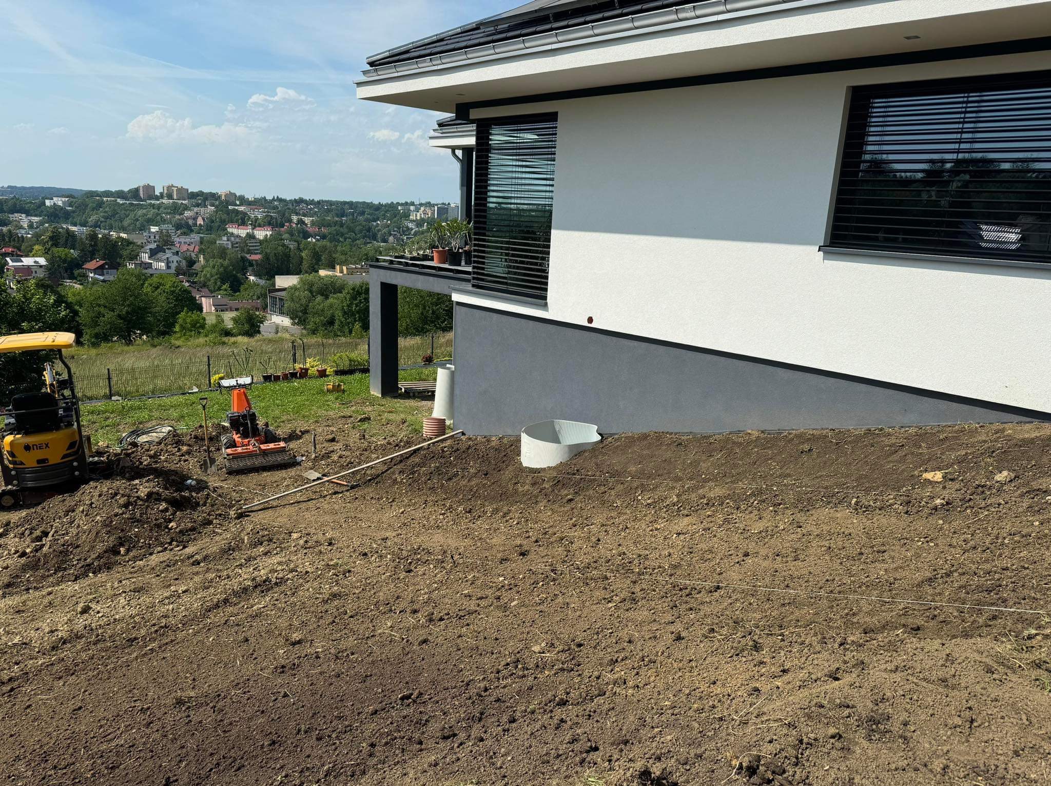 Dwa małe traktory na nierównym, przygotowanym podłożu ziemnym przed nowoczesnym domem z widokiem na miasto w tle.