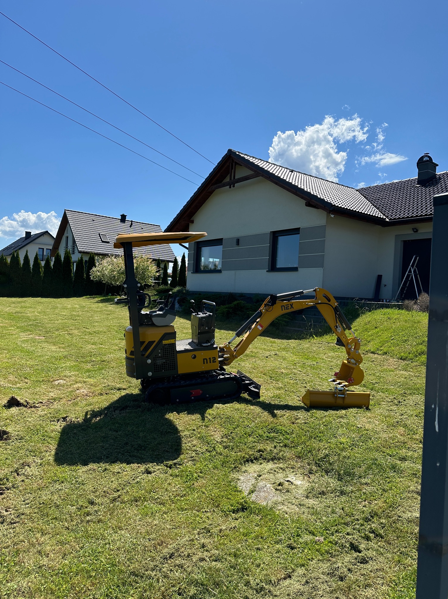 Żółta mini-koparka NEX N12 na trawniku przed domem jednorodzinnym w słoneczny dzień, widoczne ślady po pracy maszyny.
