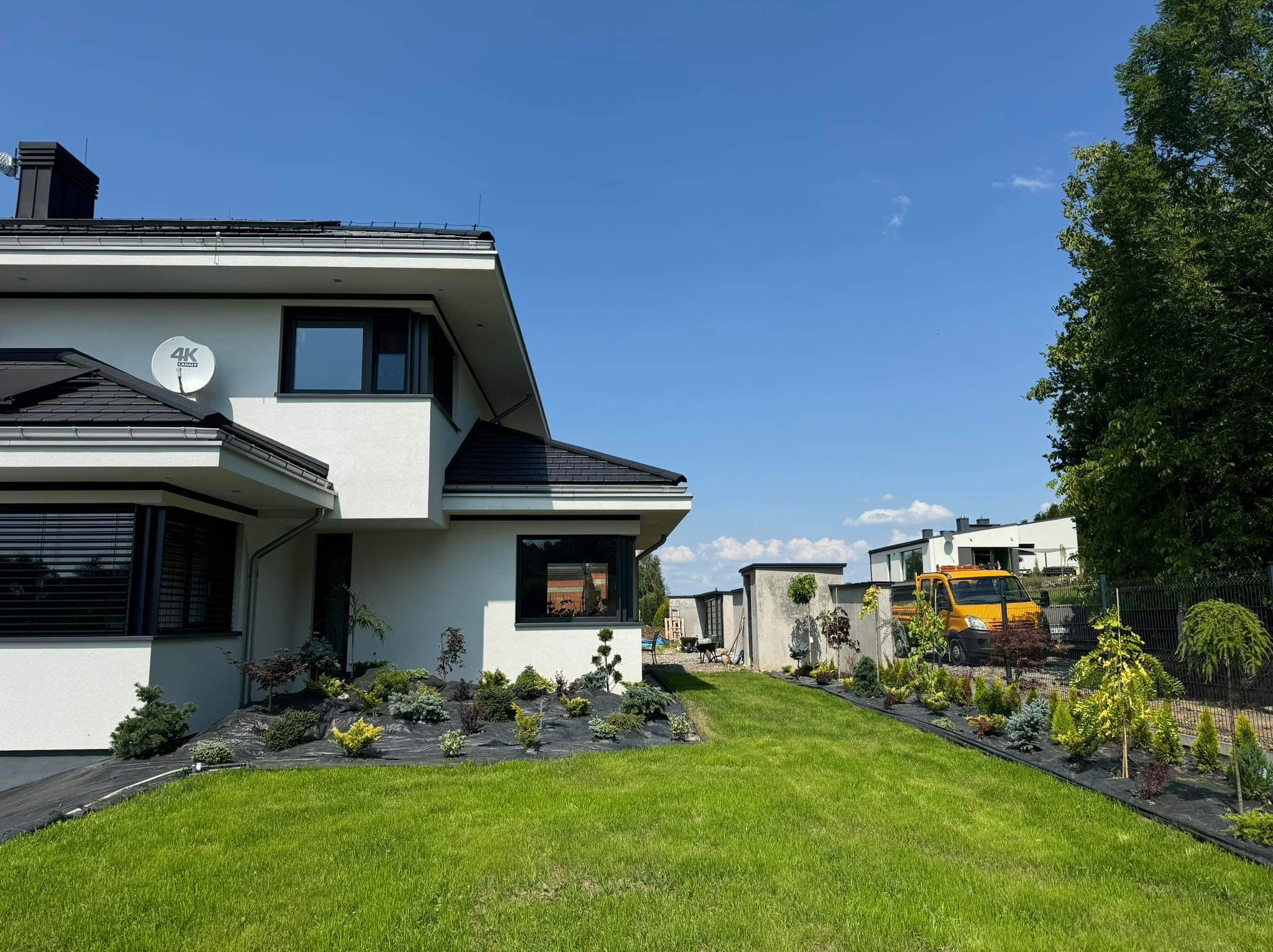 Nowoczesny dom z zagospodarowanym ogrodem: świeżo posiana trawa, rabaty z krzewami iglastymi i ozdobnymi, czarna agrowłóknina, żółty samochód dostawczy w tle.