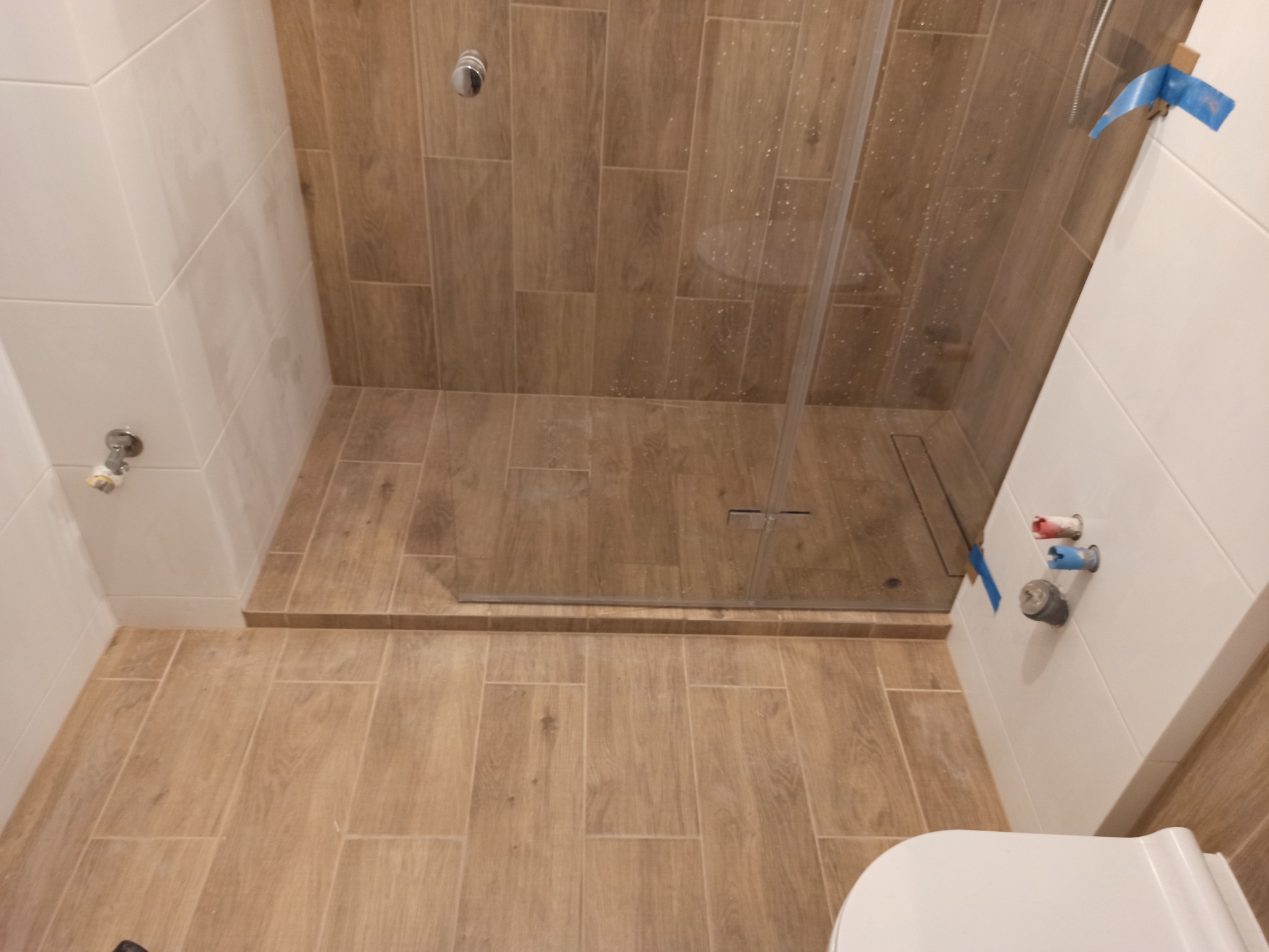 Wykończona łazienka z kabiną prysznicową typu walk-in, wyłożona płytkami imitującymi drewno, z widocznym odpływem liniowym i białą ceramiką sanitarną; widoczne elementy instalacji wodnej...