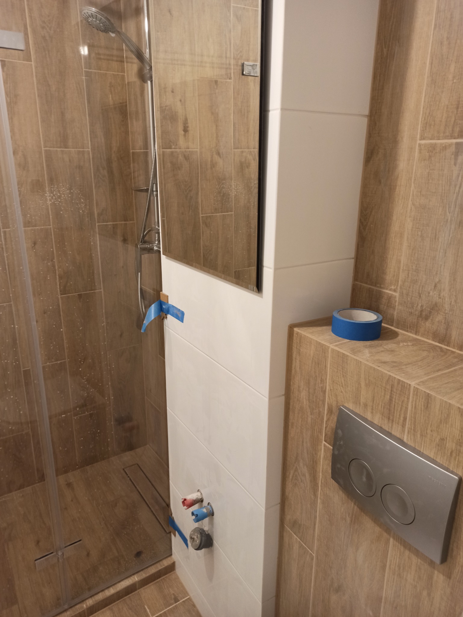 Fragment łazienki w trakcie remontu: kabina prysznicowa z odpływem liniowym, białe płytki z niebieską taśmą malarską, drewnopodobne płytki na ścianach, widoczne przyłącza wodne i przycisk spłuczki.