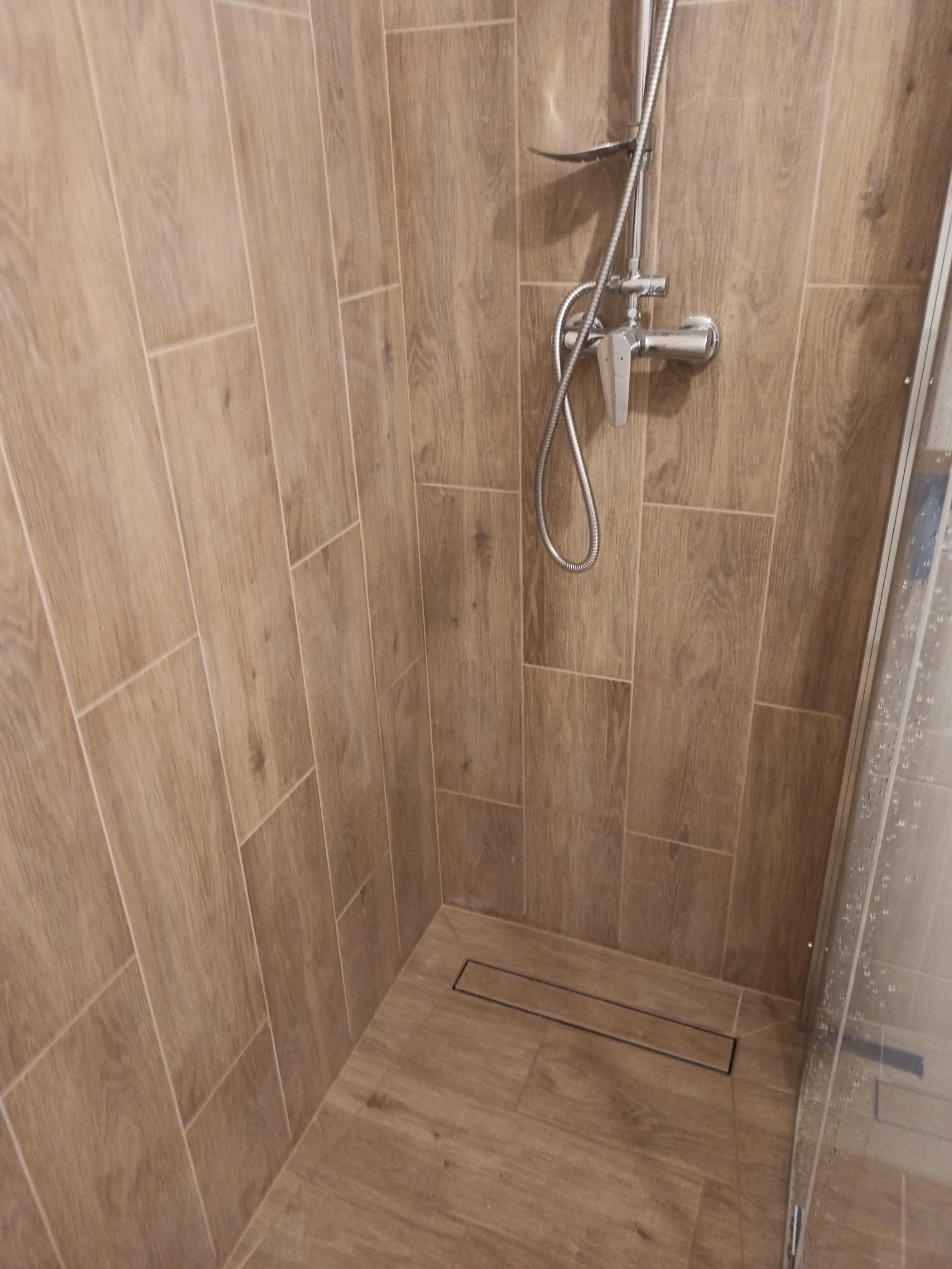 Kabina prysznicowa z deszczownicą i odpływem liniowym, wyłożona płytkami imitującymi drewno.