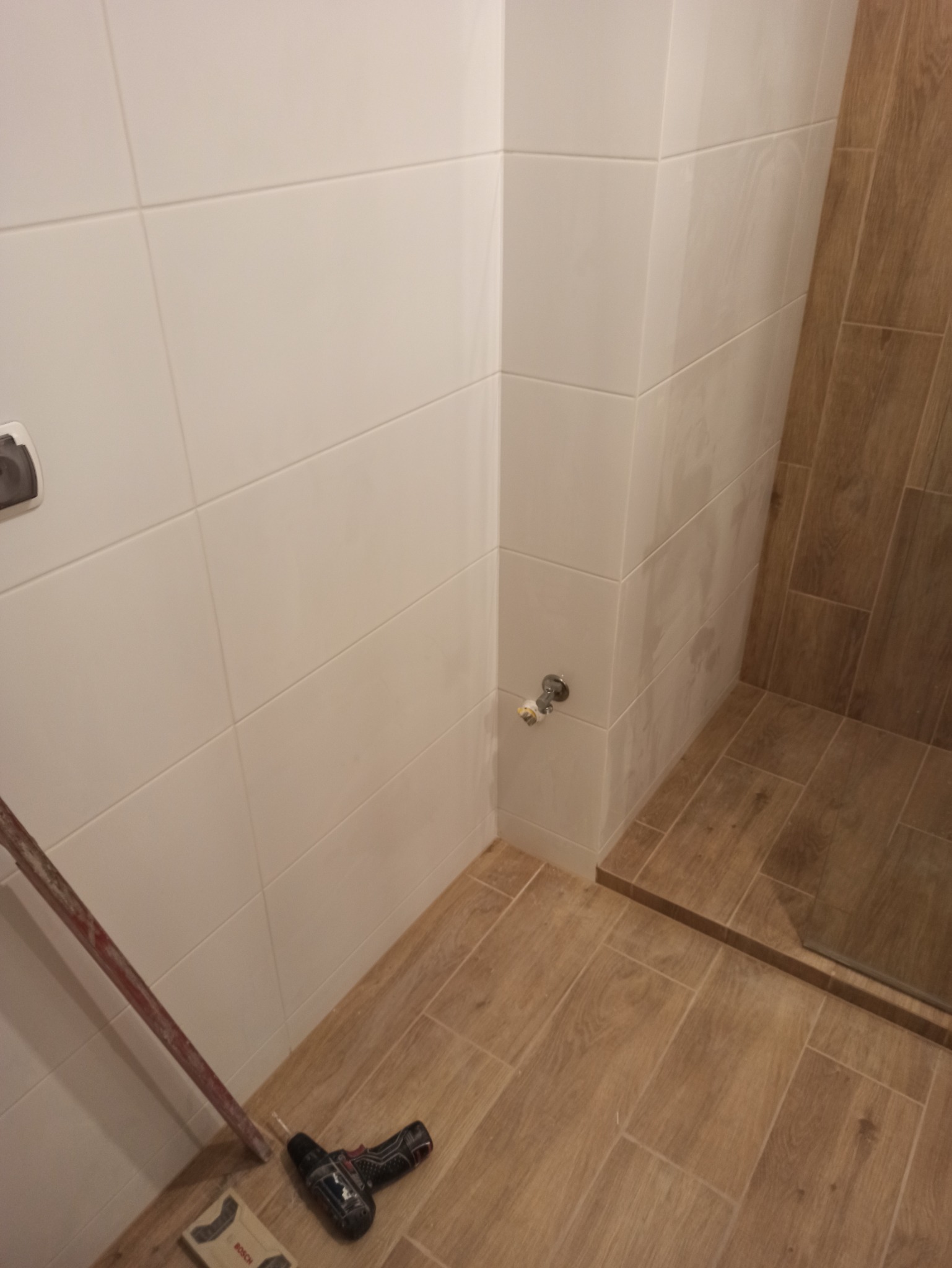 Białe, prostokątne płytki ścienne i podłogowe imitujące drewno w trakcie remontu łazienki, widoczny fragment rury, poziomica i wiertarka Bosch.