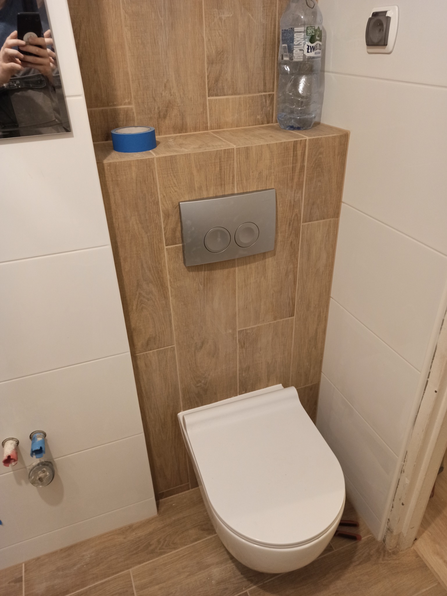 Wisząca toaleta w łazience z brązowymi płytkami imitującymi drewno, z widoczną butelką wody i niebieską taśmą malarską na półce.