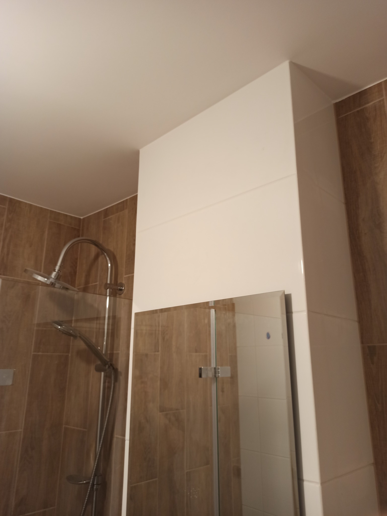 Nowoczesna kabina prysznicowa z deszczownicą, brązowe płytki imitujące drewno na ścianach, biała zabudowa z płytek na ścianie obok kabiny, lustro w kabinie.