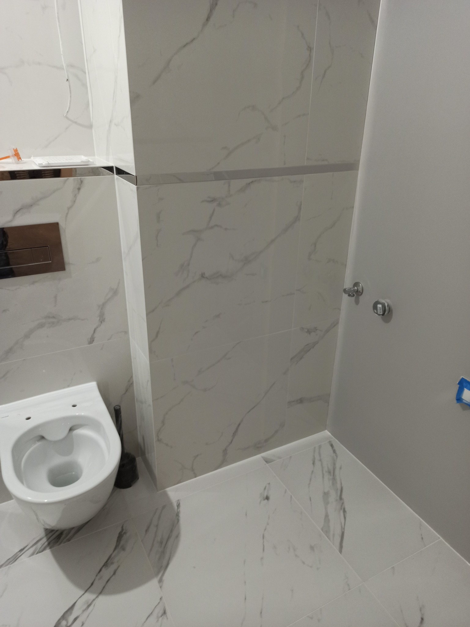 Nowoczesna łazienka z białymi płytkami imitującymi marmur, wisząca toaleta i minimalistyczna armatura, widoczne łączenia płytek z listwami.