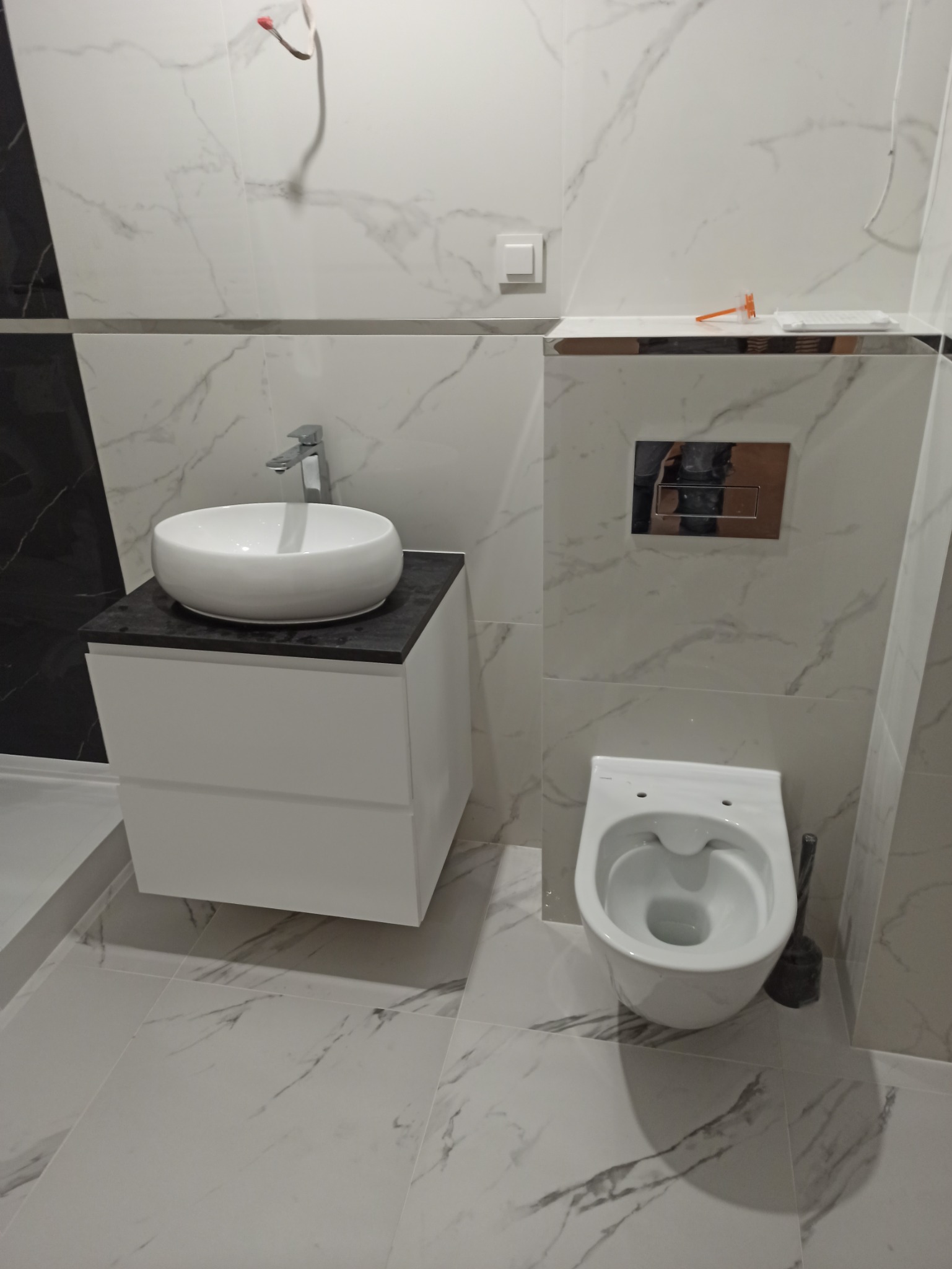 Nowoczesna łazienka w bieli i szarości: umywalka nablatowa na szafce z czarnym blatem, podwieszana toaleta, marmurowe płytki na ścianach i podłodze, widoczne elementy instalacji elektrycznej.