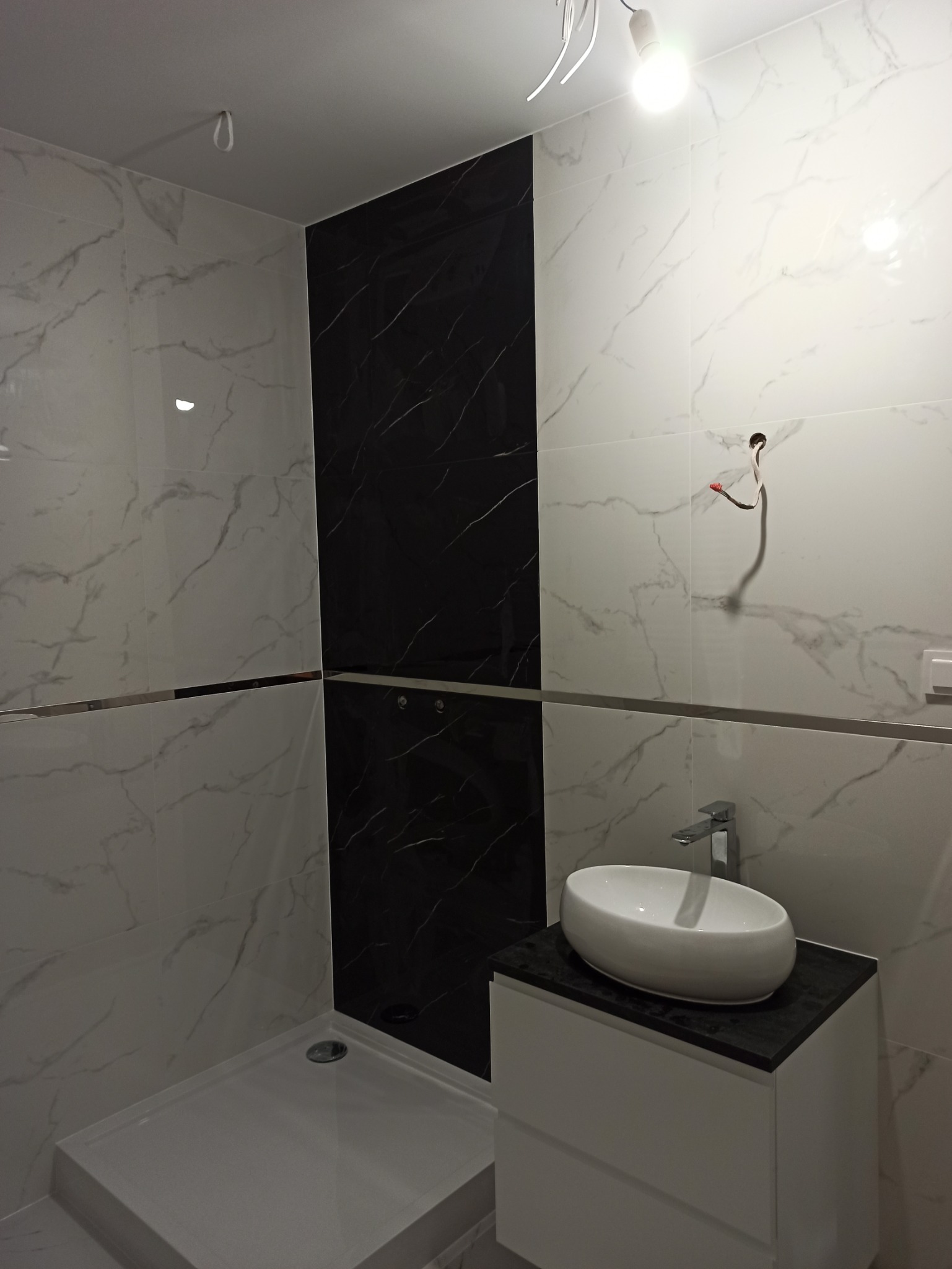 Nowoczesna łazienka w trakcie wykańczania, z czarno-białymi marmurkowymi płytkami, białą kabiną prysznicową i umywalką nablatową na białej szafce. Widoczne wystające kable elektryczne, sugerujące...
