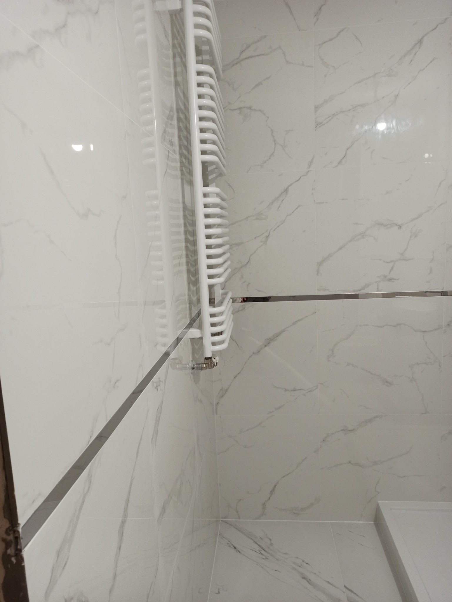 Biały grzejnik drabinkowy zamontowany na ścianie z płytek imitujących marmur w łazience, widoczna chromowana listwa wykończeniowa.