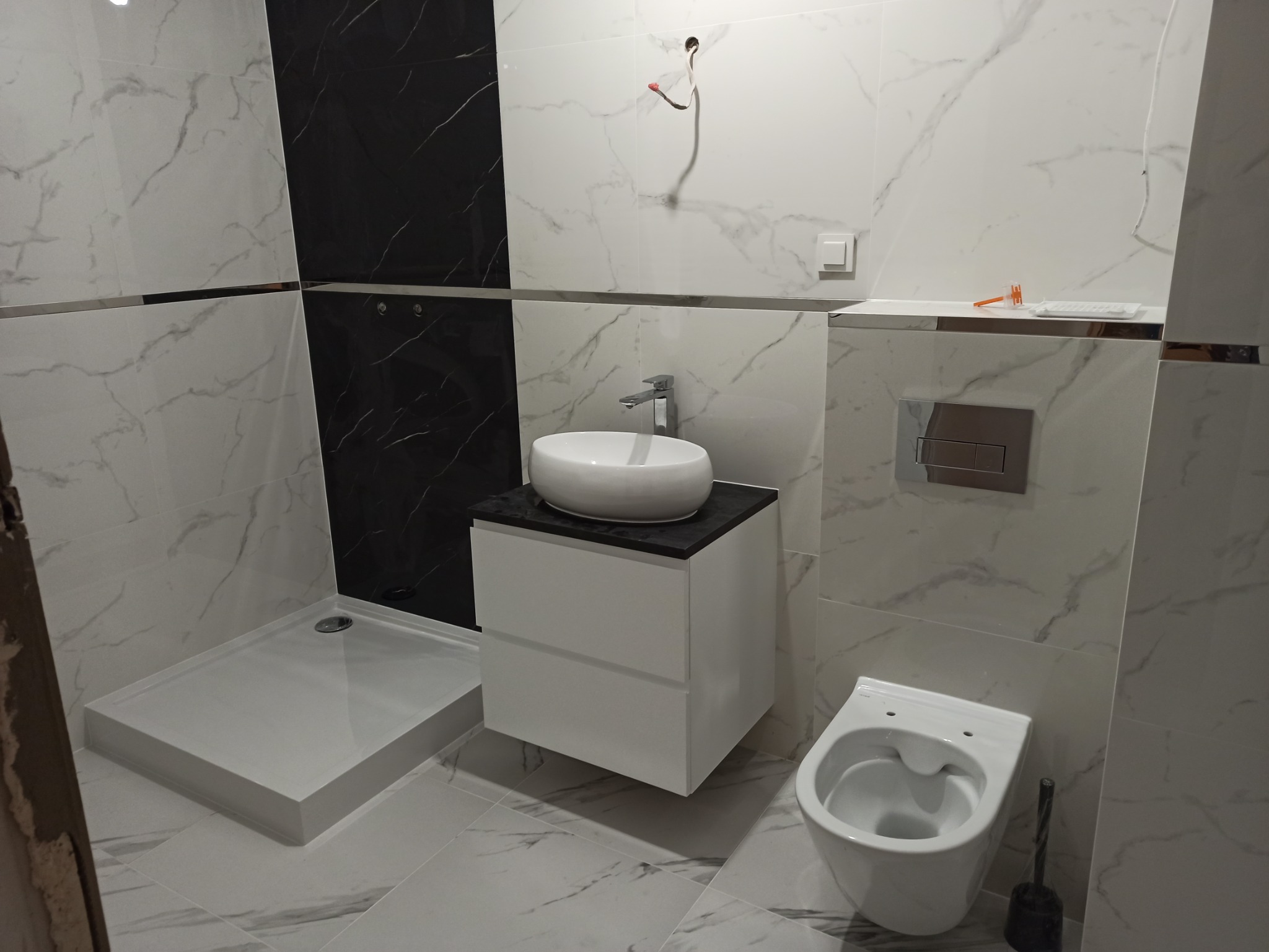 Nowoczesna łazienka z kabiną prysznicową bez brodzika, umywalką nablatową na szafce z szufladami, wiszącą toaletą i płytkami imitującymi marmur w odcieniach bieli i czerni, z widocznym fragmentem...