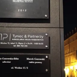 Tabliczki informacyjne na ścianie budynku w nocy: Tyniec & Partnerzy Kancelaria Usług Księgowych, Maria Gutowska-Ibbs adwokat, Marek Gutowski radca prawny, z widokiem na oświetloną ulicę.