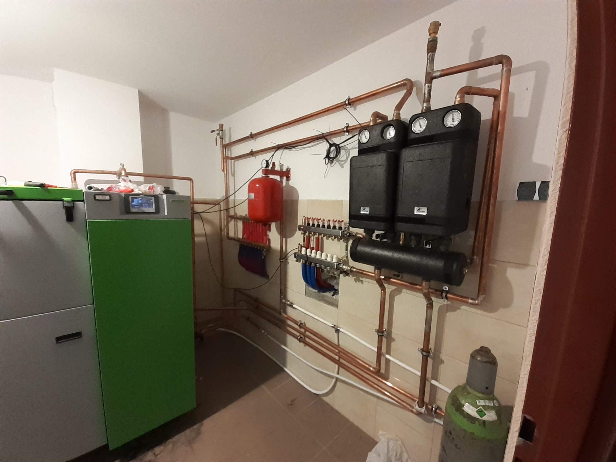 Kotłownia z nowoczesnym kotłem z zielonym panelem, dwoma czarnymi zbiornikami buforowymi z manometrami, czerwoną przeponą, rozdzielaczem hydraulicznym i miedzianą instalacją grzewczą.