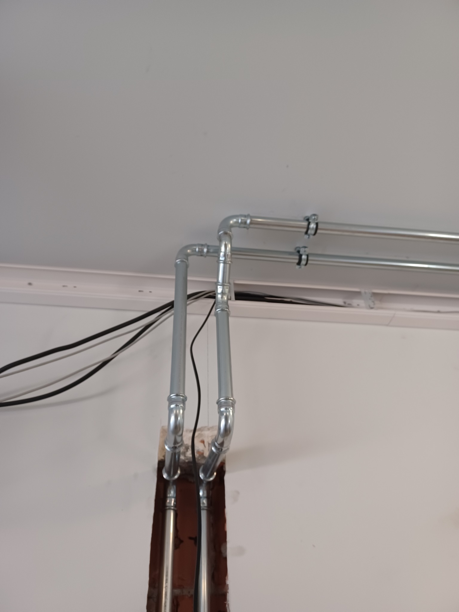 Metalowe rury instalacyjne, pionowe i poziome, przymocowane do ściany i sufitu, widoczne przewody elektryczne.