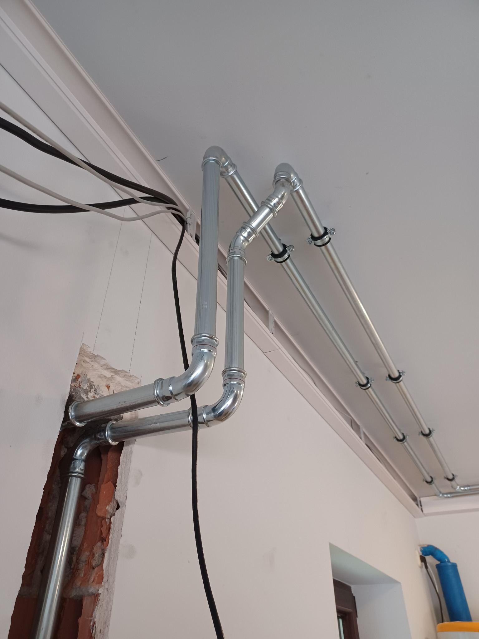 Instalacja rur gazowych/wodnych z metalu, mocowanych do sufitu za pomocą uchwytów, z widocznym fragmentem ściany z cegły w miejscu przejścia rur.