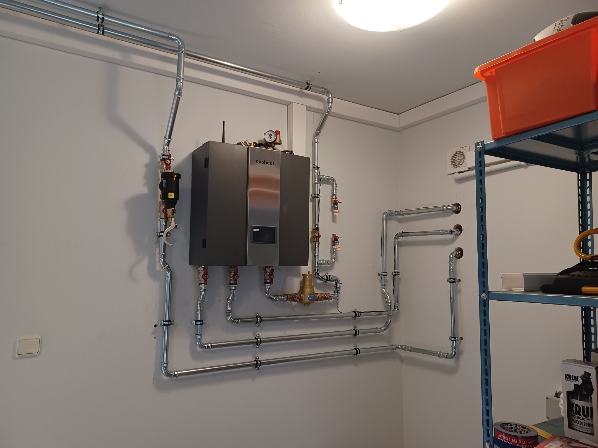 Nowoczesny, wiszący kocioł grzewczy Nesheat z kompletną instalacją rur miedzianych i zaworów, zamontowany na białej ścianie w pomieszczeniu technicznym; widoczne elementy instalacji na ścianie...