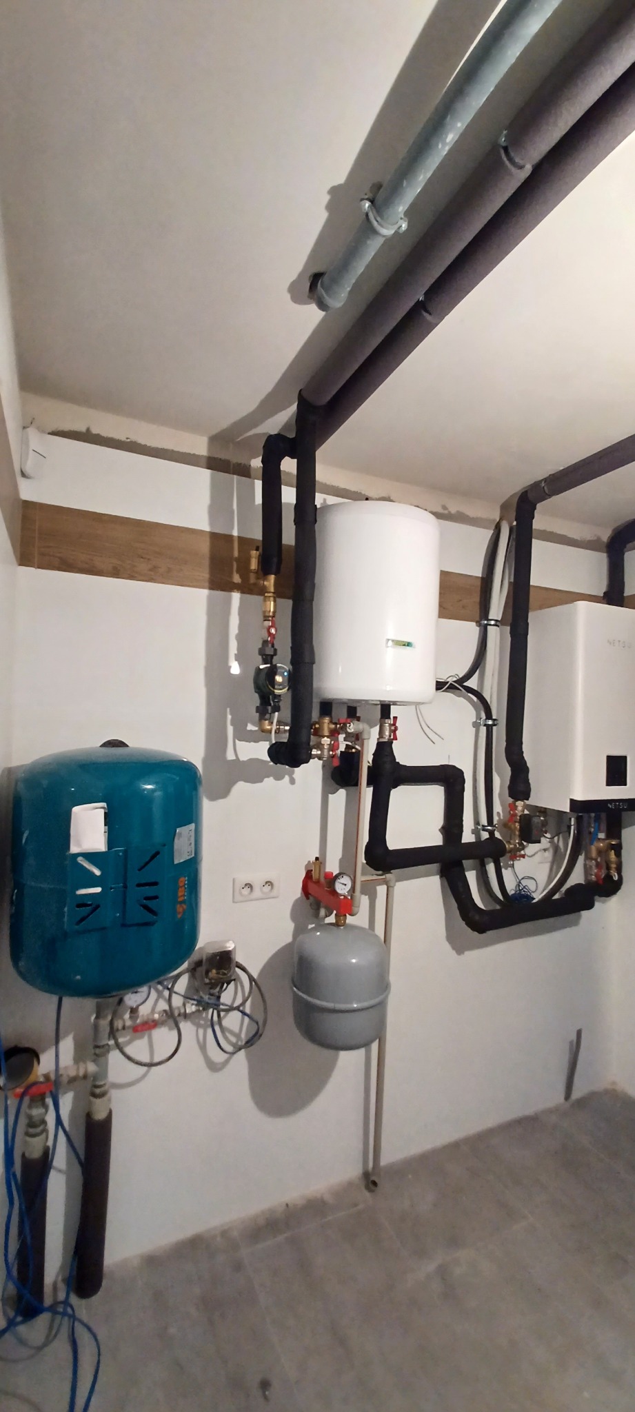 Kotłownia z zamontowanym zasobnikiem ciepłej wody, naczyniami przeponowymi, kotłem gazowym i rurami w izolacji termicznej, widok na instalację z boku.