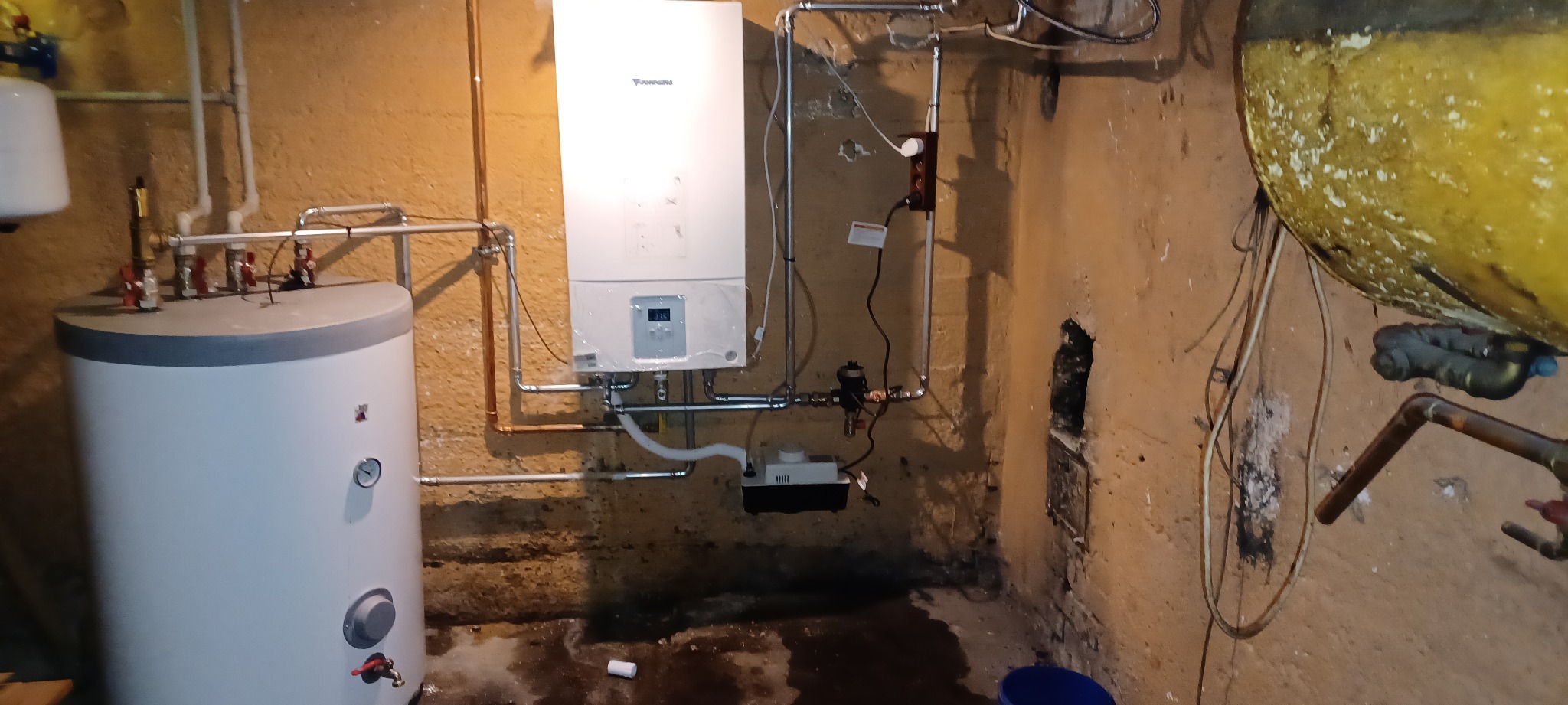 Nowy, biały, wiszący piec gazowy marki Vaillant, z podłączonymi rurami miedzianymi i plastikowymi, zamontowany w piwnicy o surowych, pomalowanych na żółto ścianach, obok biały zbiornik na wodę...