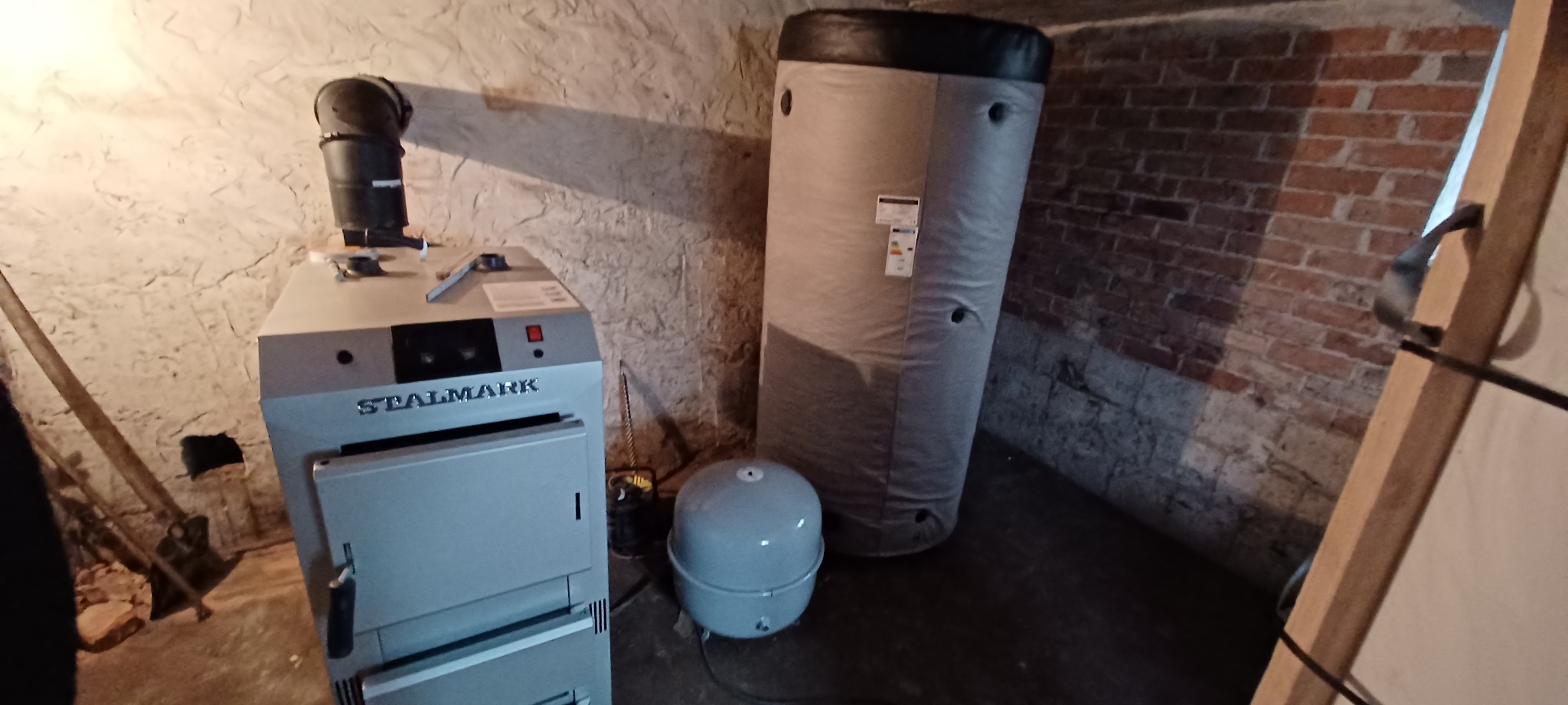 Kocioł grzewczy marki Stalmark z czarnym kominem, stojący obok szarego zbiornika akumulacyjnego i małego zbiornika wyrównawczego w piwnicy z nierównymi ścianami.