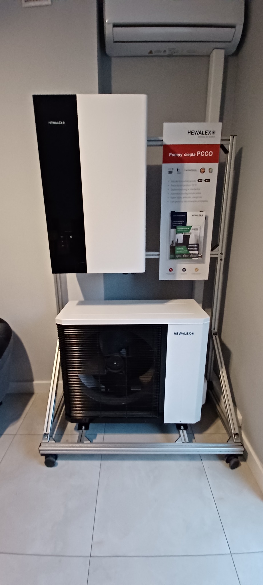 Prezentacja pompy ciepła powietrze-woda marki Hewalex na aluminiowym stojaku z kółkami, obok tablica informacyjna z danymi technicznymi, w tle klimatyzator.