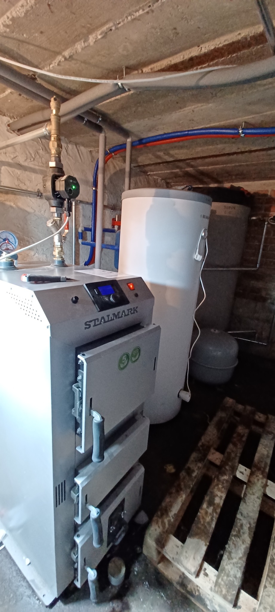 Kotłownia: nowy kocioł grzewczy marki Stalmark z otwartymi drzwiczkami, obok biały zasobnik ciepłej wody, rury instalacji grzewczej pod sufitem, podłoga na palecie.