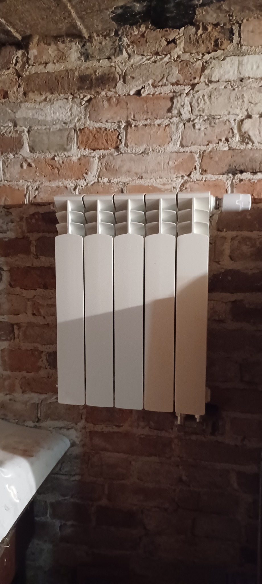 Biały, aluminiowy grzejnik panelowy zamontowany na ścianie z czerwonej, starej cegły z widocznym termostatem.