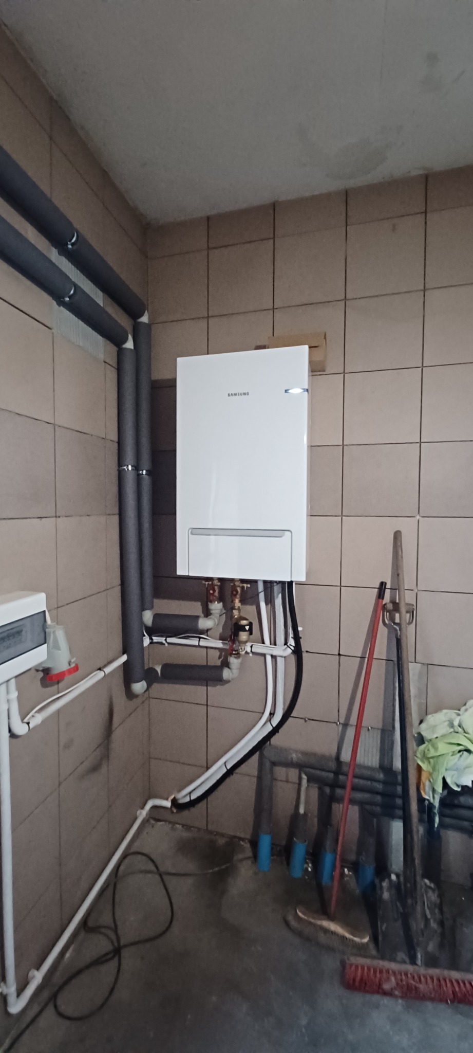 Biały, wiszący piec gazowy Samsung z podłączeniami rur miedzianych i plastikowych w pomieszczeniu z beżowymi płytkami, widoczne zaizolowane rury i akcesoria do sprzątania.