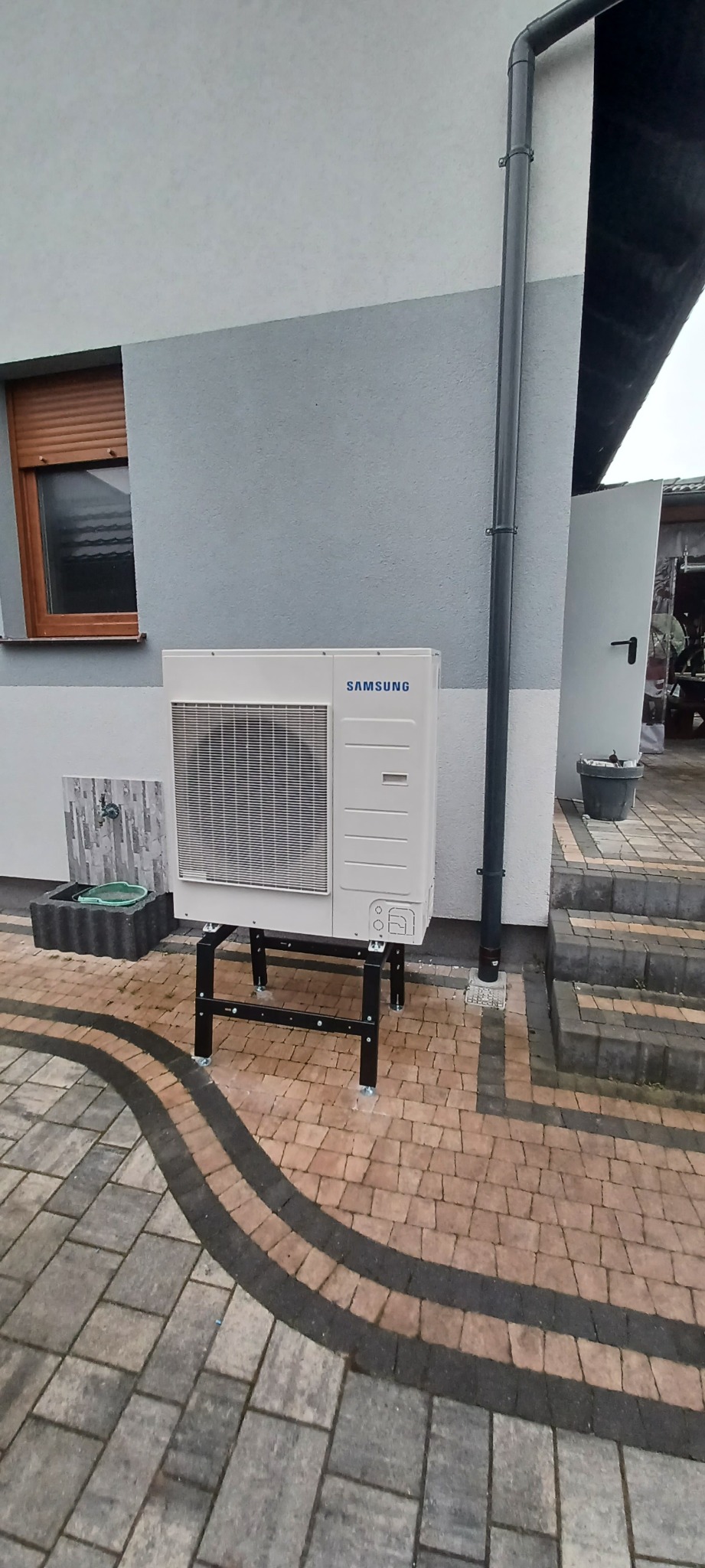 Biała zewnętrzna jednostka pompy ciepła Samsung na czarnym metalowym stojaku, ustawiona na kostce brukowej przed szarą ścianą budynku z oknem z brązowymi roletami.