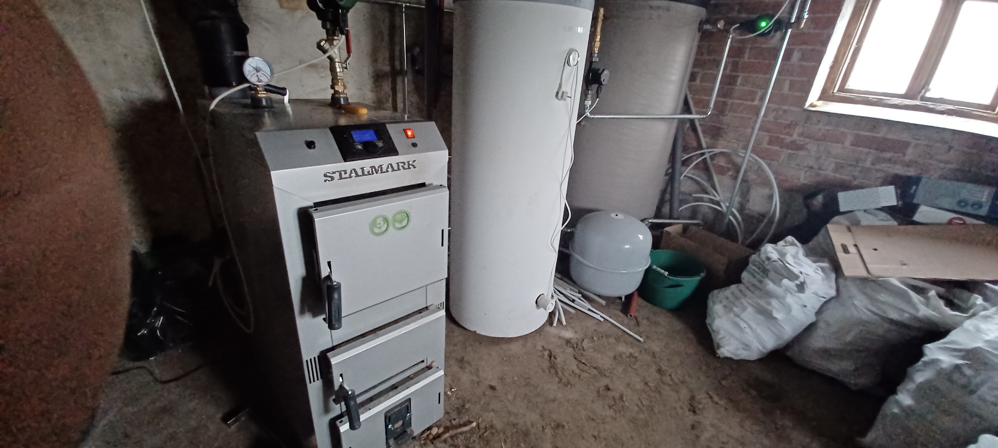 Kotłownia z kotłem marki Stalmark, zbiornikiem akumulacyjnym i osprzętem, widoczne liczniki i rury, w tle ceglana ściana i okno
