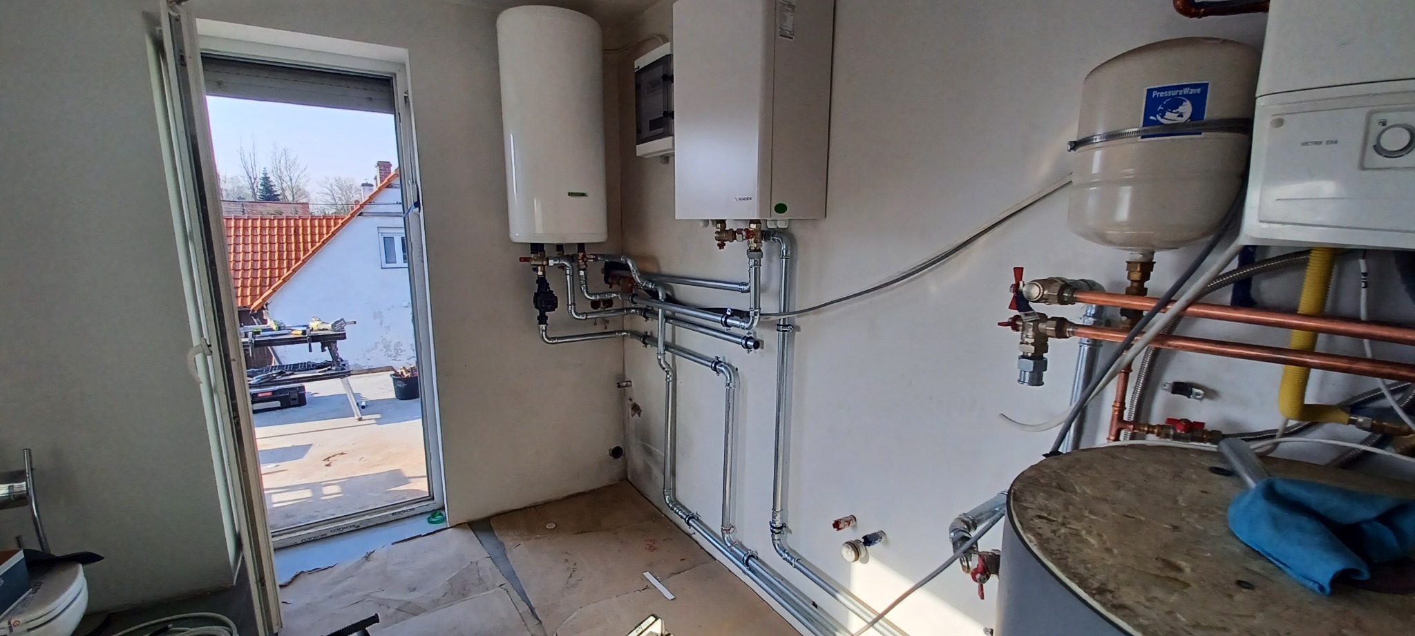 Wnętrze kotłowni w trakcie montażu: biały zasobnik ciepłej wody, wiszący kocioł gazowy, zbiornik wyrównawczy ciśnienia i instalacja rurowa z zaworami, widok na zewnątrz przez otwarte drzwi balkonowe.