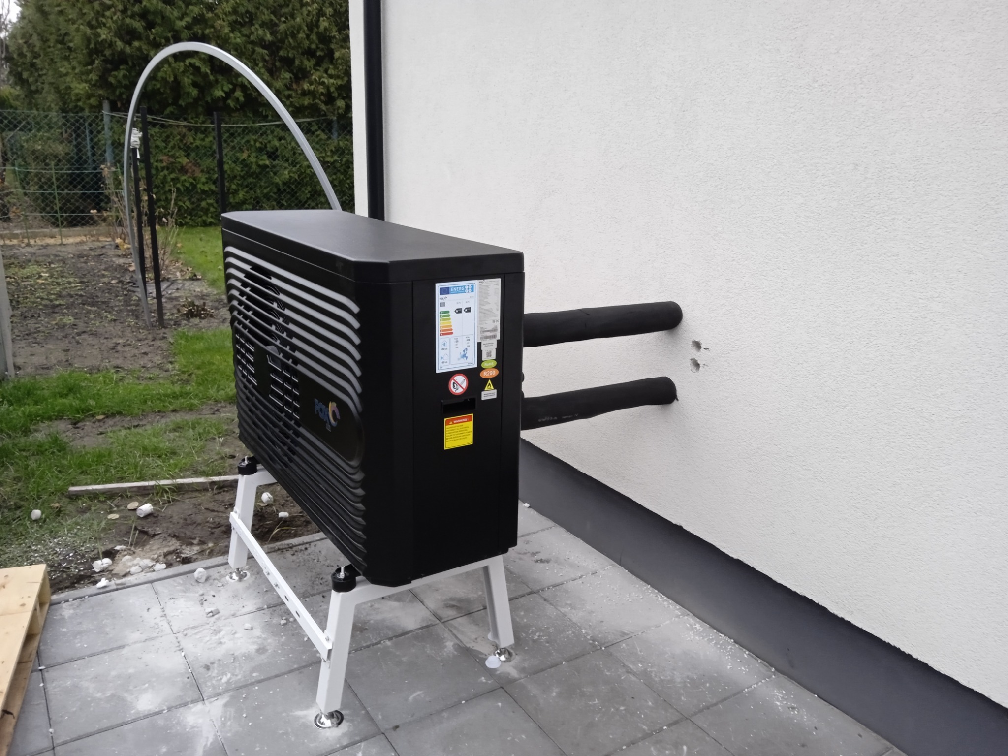 Czarna pompa ciepła powietrze-woda zamontowana na białym stelażu przy białej ścianie budynku, z widocznymi rurami w izolacji wchodzącymi w ścianę.