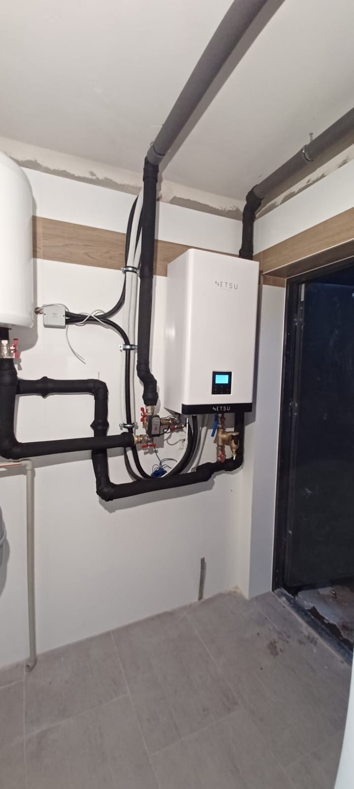 Nowoczesny, biały piec gazowy NETSU zamontowany na ścianie z widocznymi rurami instalacji grzewczej i wodnej, szare rury na suficie, świeżo wykończone wnętrze z płytkami na podłodze.