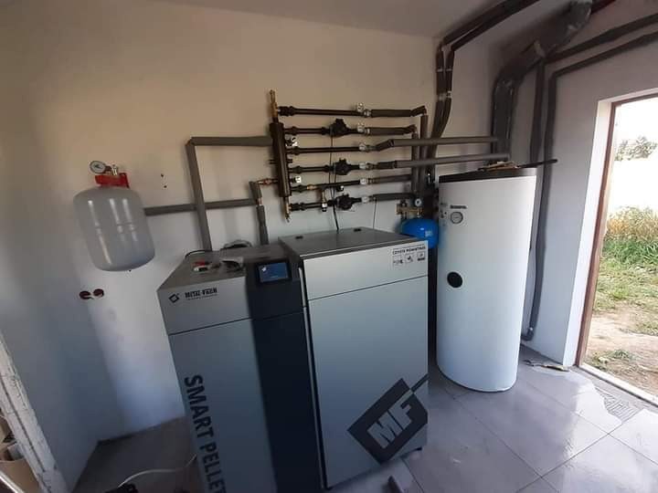 Kotłownia z kotłem na pellet, zbiornikiem akumulacyjnym i rozbudowaną instalacją rurową, widoczne elementy zabezpieczające i osprzęt.
