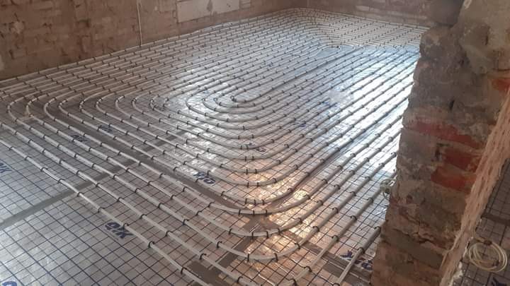 Rozłożone rury ogrzewania podłogowego na izolacji z siatką w trakcie montażu, widoczny fragment ceglanej ściany w surowym stanie.