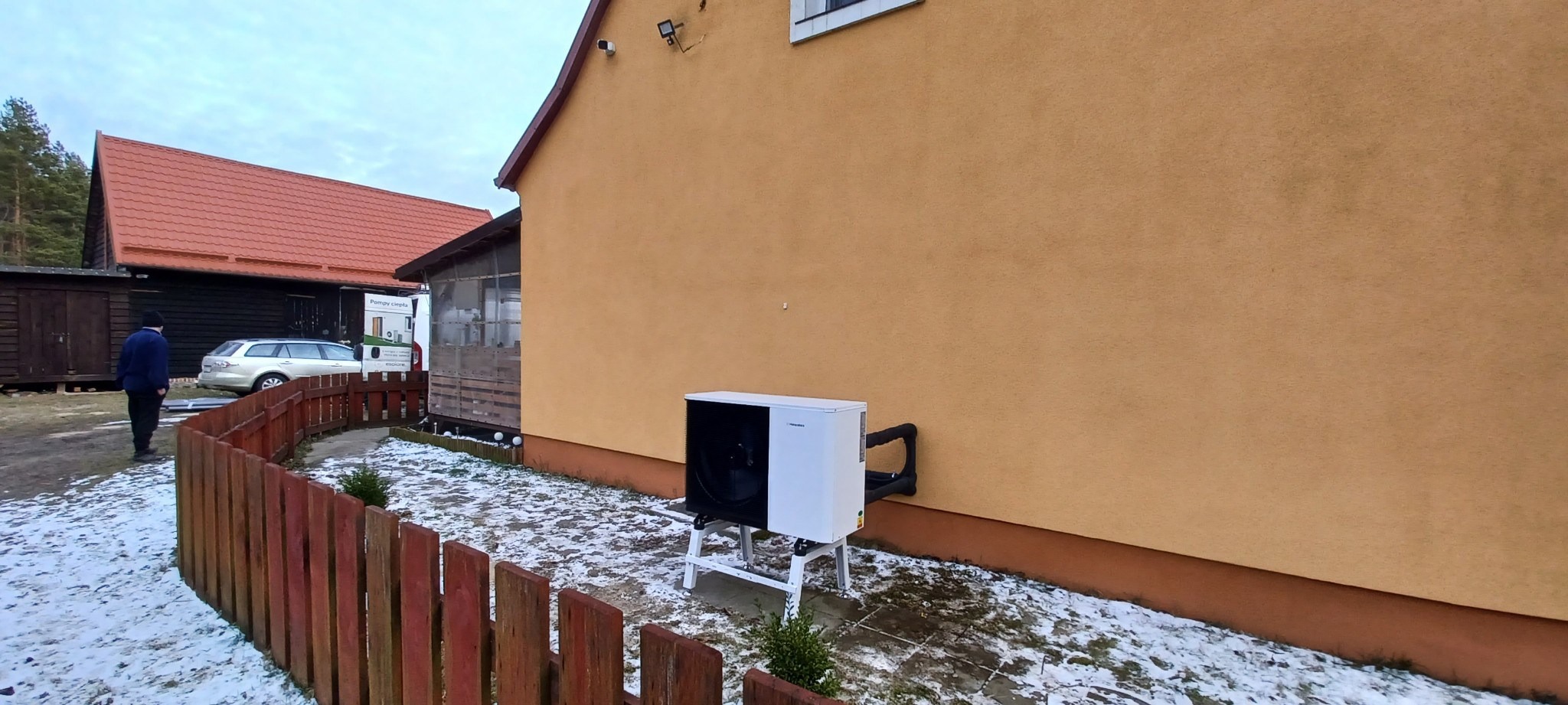 Biała pompa ciepła na metalowym stelażu, zamontowana przy żółtej ścianie domu, widoczna zza niskiego, drewnianego płotu w zimowej scenerii.