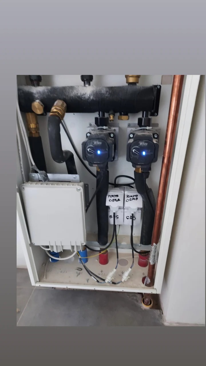Węzeł cieplny z dwoma pompami obiegowymi, orurowaniem i skrzynką sterowniczą w białej obudowie, widoczne przyłącza elektryczne i hydrauliczne.