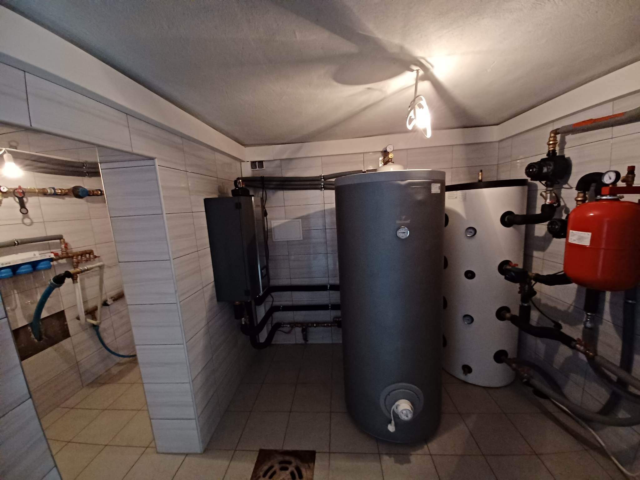 Kotłownia z dwoma zbiornikami akumulacyjnymi, piecem i instalacją hydrauliczną w pomieszczeniu wyłożonym płytkami.