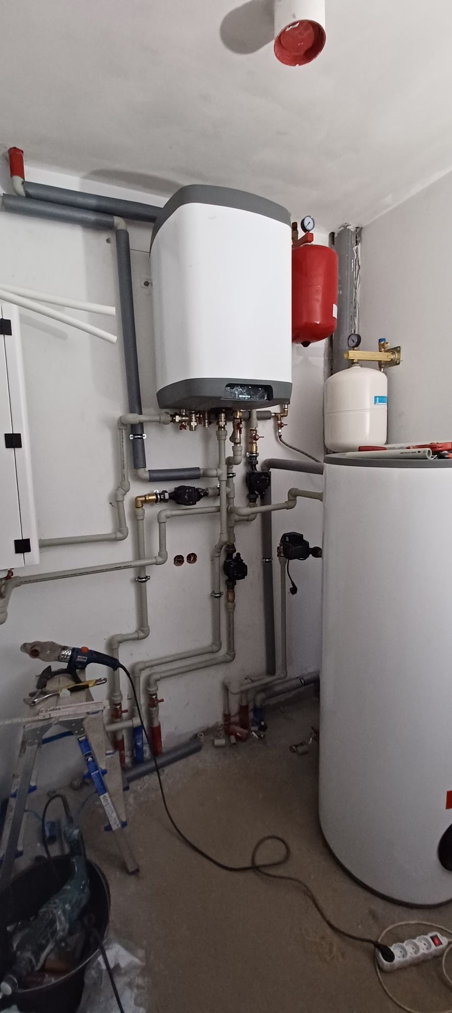Montaż kotła grzewczego z podłączonymi rurami, widoczne narzędzia na drabinie, w tle czerwony zbiornik wyrównawczy i biały zasobnik ciepłej wody.