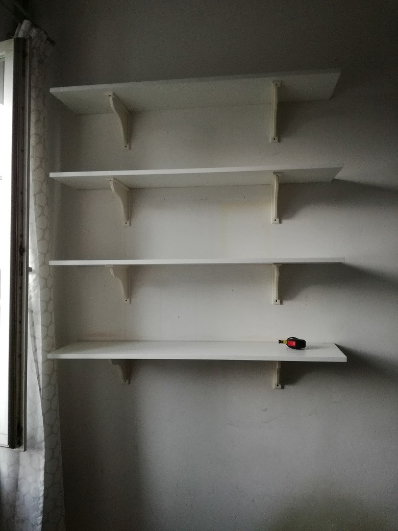 Cztery białe, puste półki zamontowane na ścianie, na najniższej półce leży zwinięta miarka.