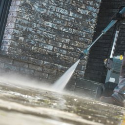 CrystalShine - Pracownik w kombinezonie roboczym czyści kostkę brukową za pomocą myjki ciśnieniowej, widoczny fragment elewacji z szarej cegły i okno.
