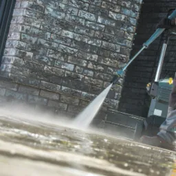 Pracownik w kombinezonie roboczym czyści kostkę brukową za pomocą myjki ciśnieniowej, widoczny fragment elewacji z szarej cegły i okno.